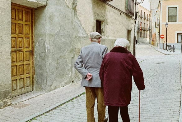 Older couple walking along a narrow cobbled street in a historic neighbourhood, representing themes of frailty and ethnicity, highlighting diverse aging experiences.