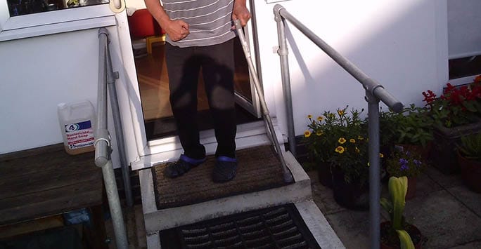 People's feet and legs, showing a person in casual clothing stepping out of a building with a wheelchair ramp, surrounded by potted plants and flowers.