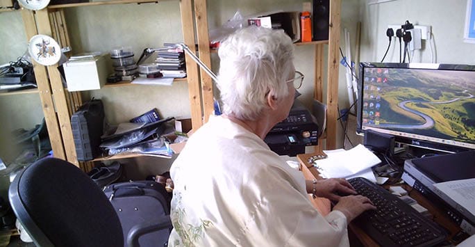 Older woman working at a desk with a computer, surrounded by books and office supplies, in a home or small study setting.