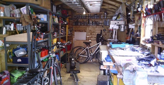 Bicycle and gym equipment in cluttered workshop or garage space filled with tools, shelves, and storage items within a wooden shed or workshop setting.
