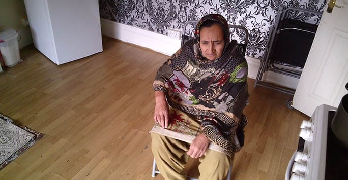 Older woman sitting in a chair, experiencing frailty, in a home setting with patterned wallpaper and wooden floor, highlighting issues of ageing and ethnicity in frailty care.