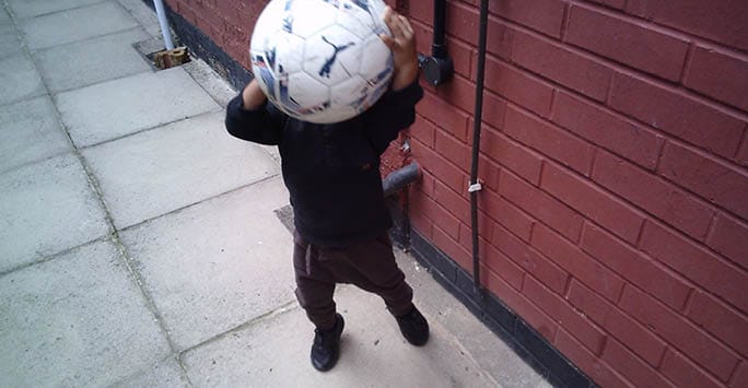 Soccer-playing child holding a ball on a sidewalk near a red brick wall, showcasing themes of physical activity and youth development in diverse communities.