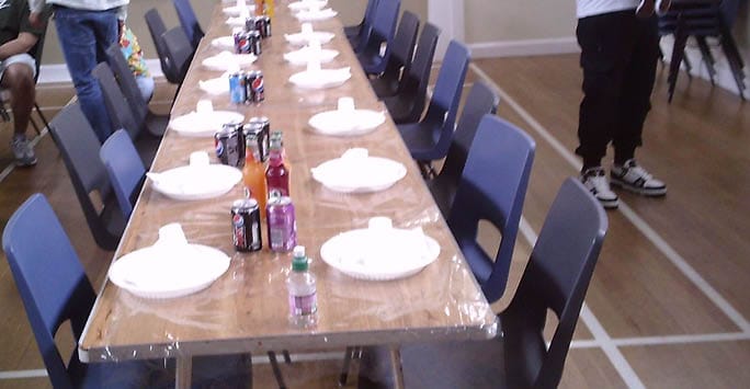A long dining table set with white plates, cutlery, and cans of drinks, surrounded by dark blue chairs in a community hall with wooden flooring and minimal decor.