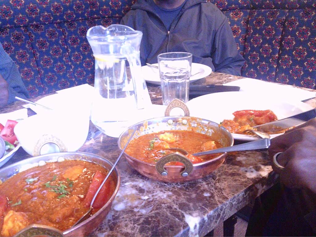 Freshly served Indian curry dishes on a marble table with water pitcher and glasses, featuring a colourful ethnic patterned wallpaper in the background.