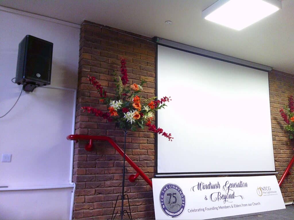 Vibrant floral arrangement next to a large white screen and a banner celebrating the founding members and elders of a church, indicating an event or presentation related to the Windrush Generation.