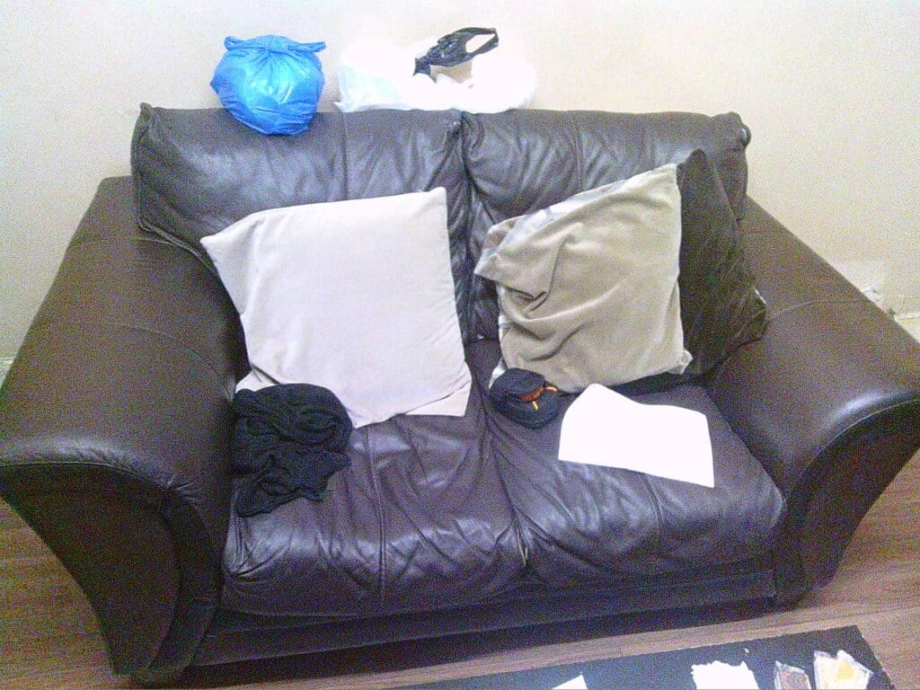 Cluttered black leather sofa with scattered clothing, pillows, and miscellaneous items, highlighting issues related to frailty and ethnicity in a domestic setting.