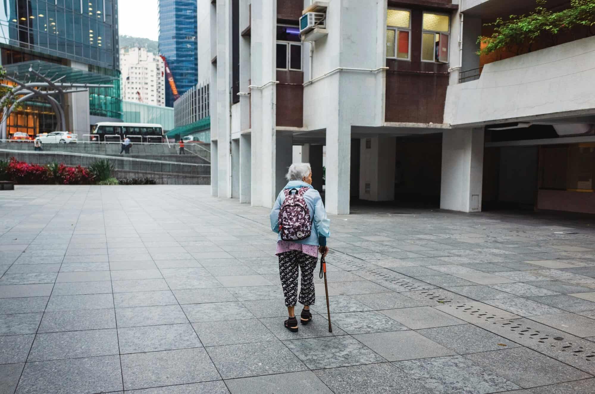 Elderly woman with a walking cane and backpack walking alone on an urban city sidewalk with modern buildings in the background.